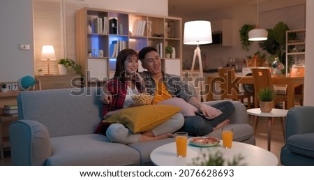 asian young couple eating pop corn are watching tv and sitting on the couch happily in night living room at home