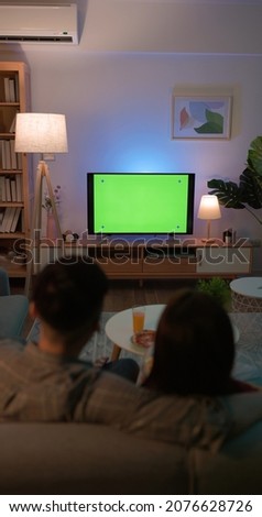 rear view of Asian young couple are watching green chroma key screen TV and having fun while sitting on couch at night in the living room