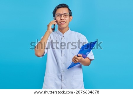 Cheerful handsome male nurse holding clipboard and talking on cell phone on yellow background