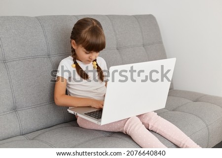 Concentrated adorable little girl with pigtail sitting on sofa in light living room playing on laptop computer or watching cartoons, typing on keyboard, looking at notebook display.