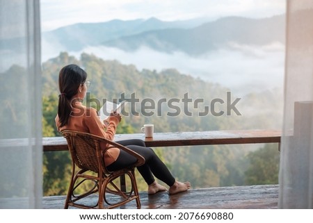 young woman reading book near window and looking mountain view at countryside homestay in the morning sunrise. SoloTravel, journey, trip and relaxing concept Royalty-Free Stock Photo #2076690880