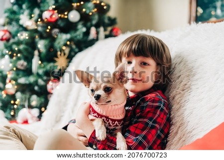 Merry Christmas. Happy little child and puppy dog in sweater playing, hug and having fun on couch with blanket at winter holidays. Kid boy in Santa hat. Christmas tree with light bokeh. Happy new year