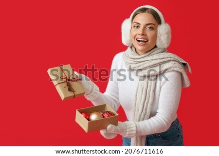 Beautiful young woman in warm earmuffs opening Christmas gift on red background