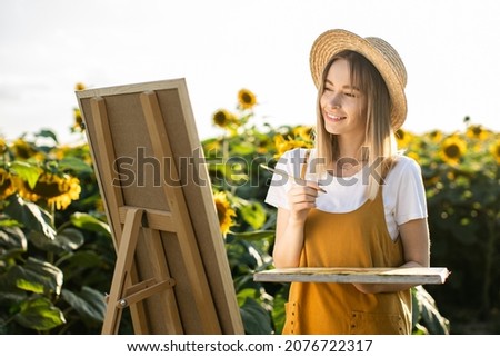 A woman is standing in a field of sunflowers and drawing a picture. The canvas is standing on an easel. She is smiling and looking at the picture. She is holding a brush and palette.