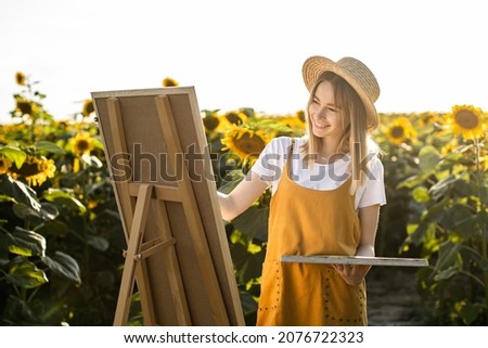 A woman is standing in a field of sunflowers and drawing a picture. She's wearing a straw hat. The canvas is standing on an easel. She is smiling. The sun is shining brightly.