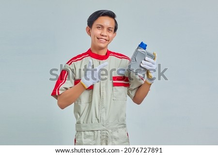 Portrait of smiling handsome man wearing mechanic uniform showing engine oil plastic bottle over gray background