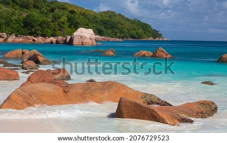 Orange stones in blue water of the Indian Ocean, white sand and green rocky hills in the background. Praslin, the Seychelles