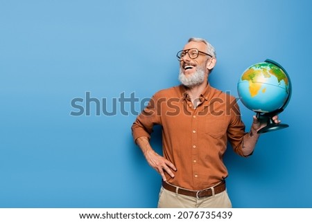 happy middle aged teacher in glasses holding globe and standing with hand on hip on blue