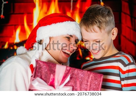 Mom in a New Year's hat gives her son in a colored sweater a Christmas present against the background of the fireplace.