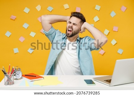 Young indignant angry successful employee business man in shirt sit work at white office desk with pc laptop put hand behind neck head look aside scream isolated on yellow background studio portrait