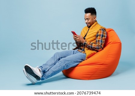 Full size body length fun young black man 20s years old wears yellow waistcoat shirt sit in bag chair hold using mobile cell phone typing isolated on plain pastel light blue background studio portrait