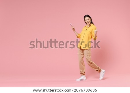 Full length side view of young brunette nice attractive latin woman 20s wearing yellow shirt hold mobile cell phone do winner gesture clench fist isolated on pastel pink background studio portrait