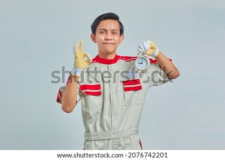 Portrait of cheerful handsome man wearing mechanic uniform holding alarm clock and showing okay sign on gray background