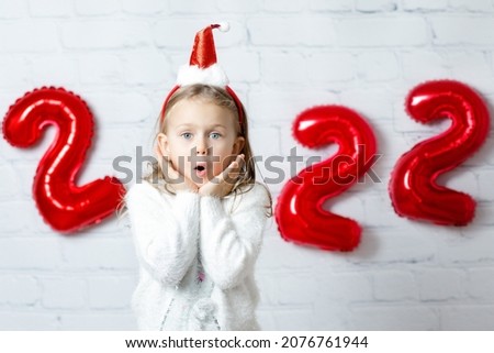 Surprised exi happy excited positive child girl in Santa hat pompon over white break wall background with figures red balloons new year numbers 2022