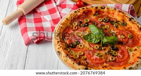 Italian pizza with pepperoni, tomatoes, olives and basil on white wooden table. Traditional Italian pizza concept. Red plaid tablecloth, pizza ingredients.