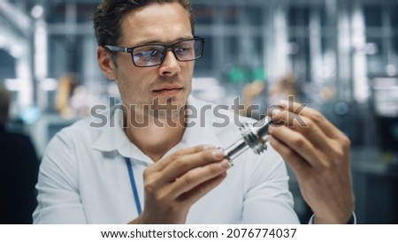 Close Up Portrait of Young Handsome Engineer in Glasses Working on Manufacturing Metal Parts in Office at Car Assembly Plant. Industrial Product Designer Examining Prototype Parts Before Production. Royalty-Free Stock Photo #2076774037