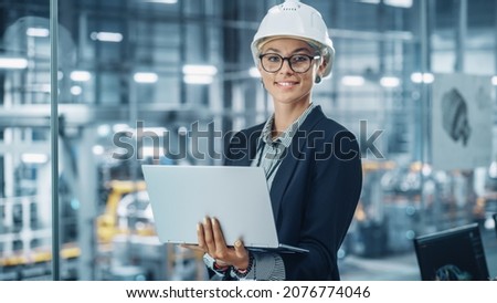 Portrait of a Happy Young Beautiful Female Engineer Wearing White Hard Hat, Using Laptop Computer in Office at Car Assembly Plant. Industrial Specialist Working on Vehicle Design in Modern Facility. Royalty-Free Stock Photo #2076774046