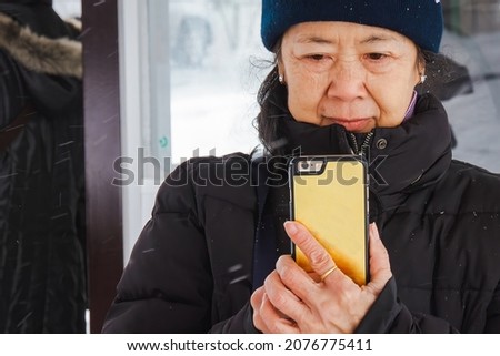 Senior Asian woman wear black winter jacket and knitted hat is smiling while take picture with smartphone camera. Mobile phone technology and winter lifestyle concept.