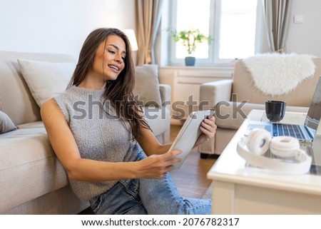Beautiful caucasian smiling woman in grey sweater and jeans sitting on sofa in living room and using tablet. Apartment interior. A young female student sitting at tfloor, using tablet when studying.