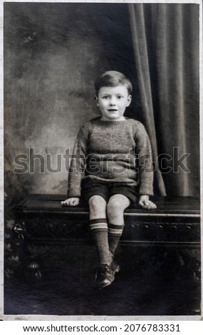 Vinatge photo of a small boy, aged 5 years. Circa 1933. Interior shot with the child seated on an ornate wooden bench, with draped curtain background.
