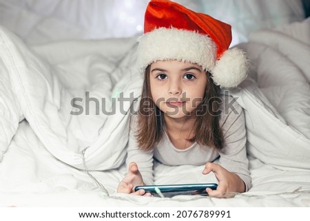 Baby little girl lying in bed in a Santa Claus hat and watching cartoons, studying, playing a game on the phone, looking at the camera, smiling