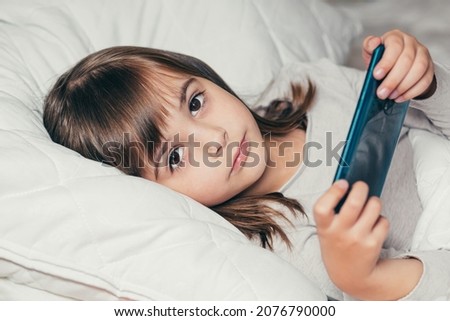 Baby little girl lying in bed with phone and watching cartoons or playing a game on the phone and smiling looking at the camera