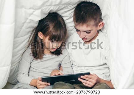 Two children a boy and a girl, a brother and a sister friends sit under a white blanket on the bed and play, read, study on the tablet