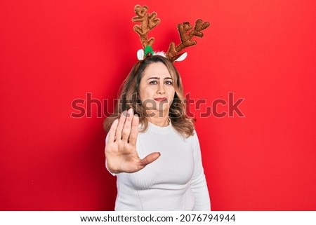 Middle age caucasian woman wearing cute christmas reindeer horns doing stop sing with palm of the hand. warning expression with negative and serious gesture on the face. 