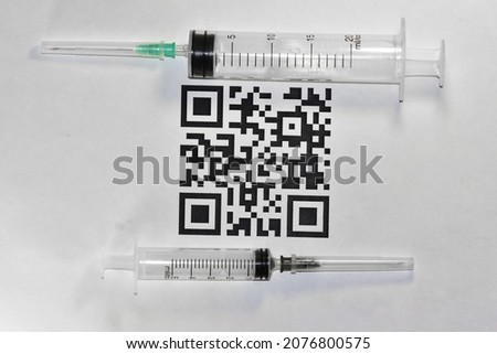A huge medical syringe lies on a sheet with the QR-code "VACCINED COVID-19"