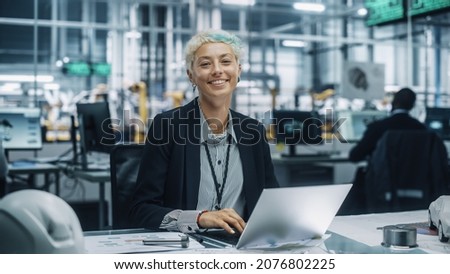 Portrait of a Happy Young Beautiful Female Engineer Sitting at a Desk, Using Laptop Computer in Office at Car Assembly Plant. Industrial Specialist Working on Vehicle Design in Modern Facility. Royalty-Free Stock Photo #2076802225