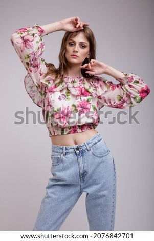 High fashion photo of a beautiful elegant young woman in a pretty blouse top with floral pattern in red, blue jeans posing over white, soft gray background. Slim figure. Studio Shot.