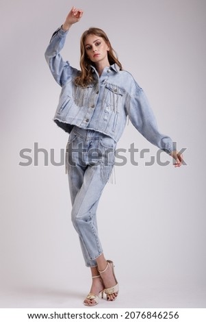 High fashion photo of a beautiful elegant young woman in a pretty denim jeans oversize jacket with rhinestones, pants posing over white, soft gray background. Slim figure.  Studio Shot.