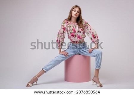 High fashion photo of a beautiful elegant young woman in a pretty blouse top with floral pattern in red, blue jeans posing over white, soft gray background. Model is sitting on a pink cube cylinder