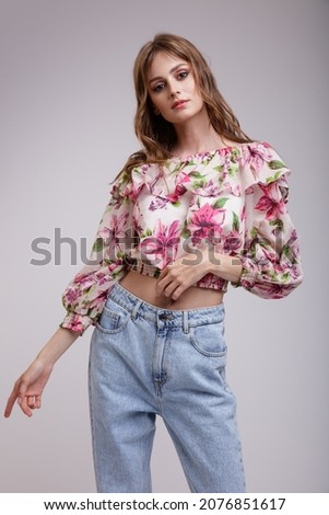 High fashion photo of a beautiful elegant young woman in a pretty blouse top with floral pattern in red, blue jeans posing over white, soft gray background. Slim figure. Studio Shot.