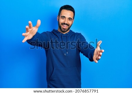 Handsome hispanic man with beard wearing casual sweatshirt looking at the camera smiling with open arms for hug. cheerful expression embracing happiness.  Royalty-Free Stock Photo #2076857791