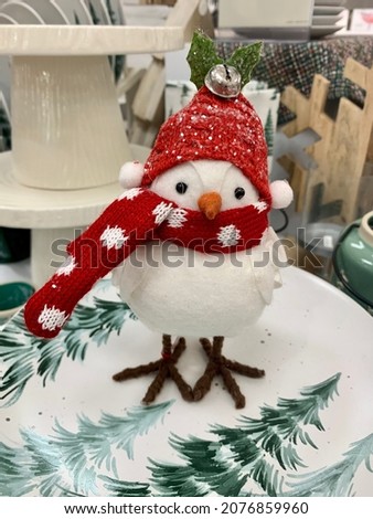 The top down, close up image of a decorative bird wearing a red scarf and hat. The penguin figurine is sitting on a festive plate with green pine trees and next to a green mug with a spoon attached.