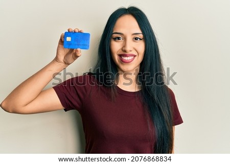 Young hispanic girl holding credit card looking positive and happy standing and smiling with a confident smile showing teeth 