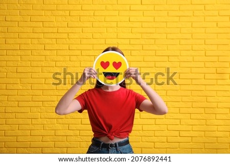 Woman covering face with heart eyes emoji near yellow brick wall Royalty-Free Stock Photo #2076892441