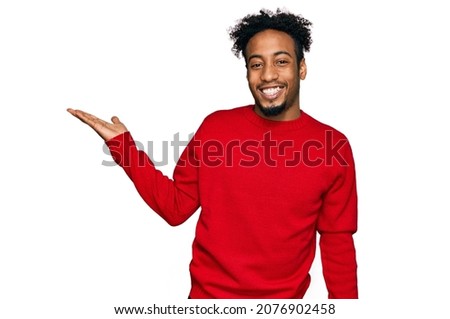 Young african american man with beard wearing casual winter sweater smiling cheerful presenting and pointing with palm of hand looking at the camera. 