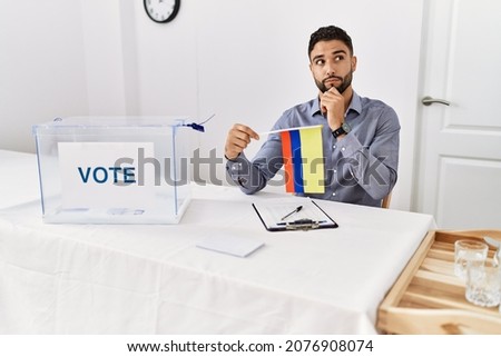 Young handsome man with beard at political campaign election holding colombia flag serious face thinking about question with hand on chin, thoughtful about confusing idea 