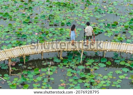 Top view of Asian tourists enjoy taking pictures on bamboo bridge over river with many lotuses. Happiness couple spending time together outside in water nature. Scenery view of people with lotus pond.