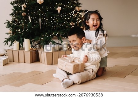 Funny happy Asian kids brother and sister hugging giving gift boxes celebrating Christmas holiday sitting at the decorated Christmas tree at home. Selective focus