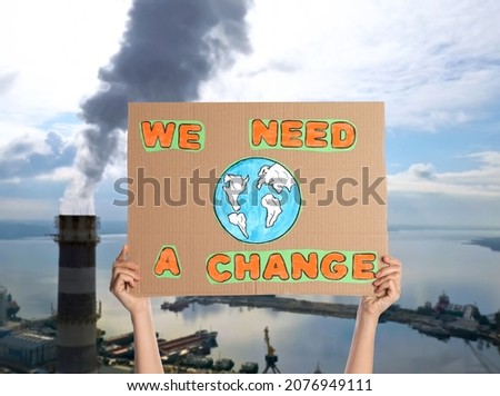 Protestor holding placard with text We Need A Change and blurred view of industrial factory on background. Climate strike