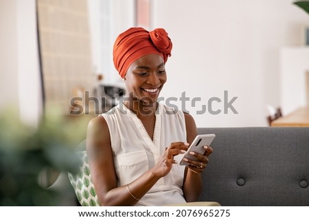 Smiling middle aged african woman with traditional head turban sitting on couch at home using smartphone. Beautiful african american woman with typical headscarf scrolling through internet on phone. Royalty-Free Stock Photo #2076965275