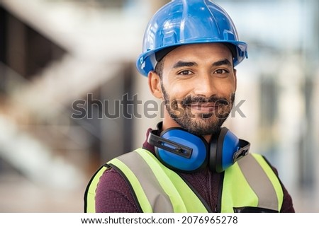 Portrait of satisfied construction site manager wearing safety vest and blue helmet with copy space. Young middle eastern architect watching construction site with confidence and looking at camera. Royalty-Free Stock Photo #2076965278