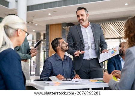 Successful entrepreneur focusing on work with charts during meeting with multiethnic employee. Mature businessman discussing work at conference table in office with senior businesswoman.