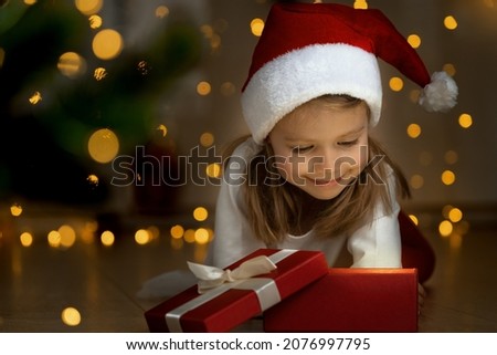 Little girl in red Santa Claus hat opens box with light at dark next to christmas tree. Happy kid with New Year gift