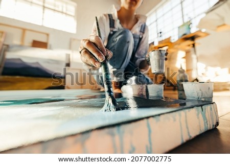 Unrecognizable female artist painting on a large canvas with a paintbrush. Young painter working on the floor of her art studio. Creative young woman making a blue painting for her new art project. Royalty-Free Stock Photo #2077002775