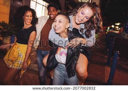 Carefree young woman piggybacking her friend in the city. Girlfriends having a good time while going out with their friends at night. Group of vibrant friends hanging out together on the weekend. Royalty-Free Stock Photo #2077002832