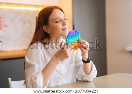 Close up face of happy charming redhead young woman pushing colorful iridescent soft silicone bubbles at home. Smiling female with trendy stress and anxiety relief fidgeting game in living room.